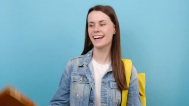 Portre mutlu genç kız parmakla işaret ediyor, baş parmağını gösteriyor, sarı sırt çantası takıyor, stüdyoda mavi arka planda yalnız poz veriyor. Lise üniversite konsepti eğitimi