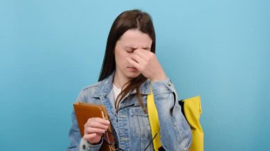 Tired girl teen student hold books glasses rub nose, smiling looking at camera, wear denim jacket and yellow backpack, isolated on blue background. Education in high school university college concept