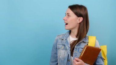 Portre gülümseyen genç kız, mavi duvarda izole edilmiş, promosyon içeriği için üzerinde sarı ok bulunan kot ceket ve sırt çantası giyiyor. Üniversite konsepti eğitimi