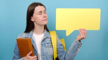 Thoughtful pensive girl student showing empty speech bubble thinking, wears denim jacket and yellow backpack, posing isolated over blue color background in studio with copy space for advertisement