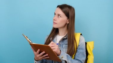 Düşünceli genç kız öğrencinin portresi kot ceket giyer ve egzersiz kitabı notlarına sarı sırt çantası yazar. Stüdyonun mavi arka plan duvarına izole edilmiştir. Üniversite üniversitesinde eğitim