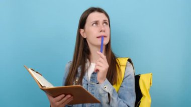 Telaşlı genç kız öğrencilerin portresi kot ceket giyer ve egzersiz kitabı notlarına sarı sırt çantası yazarak stüdyoda mavi renkli arka plan üzerinde izole bir poz verir. Üniversite üniversitesinde eğitim