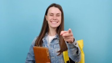 Portre mutlu genç kız kitapları göz kırpma göz kırpma, kot ceket giyiyor ve sarı sırt çantası, stüdyoda mavi renkli arka planda izole. Lise üniversite konsepti eğitimi