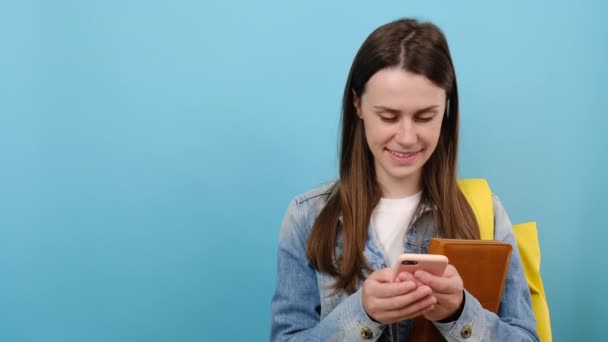 笑着的未成年女生穿着斜纹棉布夹克和黄色背包的画像用手机把书架放在工作区上模仿空间 隔离在蓝色的工作室背景墙上 — 图库视频影像