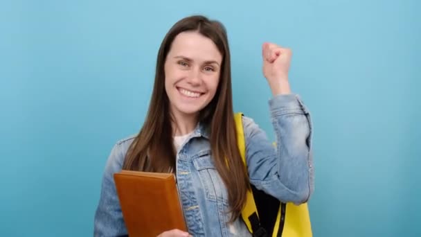 快乐可爱的女生穿着斜纹棉布夹克 黄色的背包紧握着书本 在苍白的蓝色背景工作室里显得孤立无援 高中学院教育的概念 — 图库视频影像