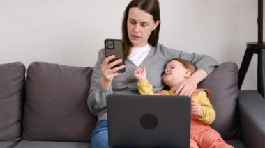 Çekici genç anne uzaktan kumandası çevrimiçi çalışıyor. Şirin küçük kızının yanında rahat bir koltukta oturuyor. Annesi dizüstü bilgisayar ve akıllı telefon kullanıyor. Modern aile araç gereç kullanır. Sanal ev ofisi, uzak iş