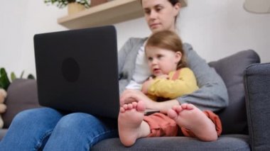 Mutlu genç annenin seçici odağı küçük kızını tutuyor, annesi dizüstü bilgisayarda klavye kullanıyor evde bebekle rahat bir yatakta oturup bilgisayar uygulamaları teknoloji konseptiyle çalışıyor.