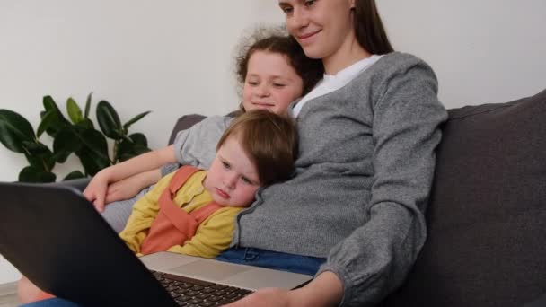 幸せな若いお母さんの近くに2人のかなり小さな娘を抱きかかえて コンピュータを使って教え 教育ビデオを見て 現代的なデバイスで楽しんでいるノートブックスクリーンを見て居心地の良いソファに座って — ストック動画