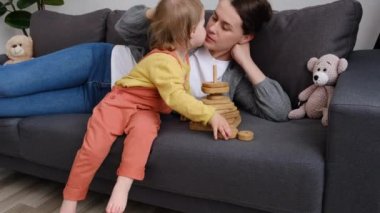 Mutlu gülümseyen genç anne, küçük tatlı kızın birlikte oyuncaklar oynamasına yardım ediyor. Güzel, şefkatli anne ve küçük kız, kahkahalar atan, ahşap bloklardan bir kule inşa ederek eğleniyorlar.