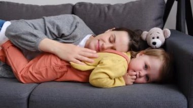 Sevecen, genç, beyaz bir annenin, tatlı, küçük kızıyla, evdeki rahat, gri kanepede uzandığı o hassas anın tadını çıkarmasına yakın. Mutlu anne, gülen küçük bebekle vakit geçiriyor.