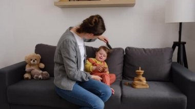 Genç, şefkatli bir anne, güzel küçük bir kızın saçlarını tarıyor. Gülümseyen anne, evdeki oturma odasında rahat bir koltukta oturan küçük kızına saç stili yapıyor. Annelik ailesi sevgisi
