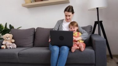 Küçük kızıyla birlikte gülümseyen genç bir anne dizüstü bilgisayarla komik videolar izliyor çevrimiçi TV izliyorlar hafta sonları evde birlikte vakit geçiriyorlar oturma odasında rahat koltukta oturuyorlar.