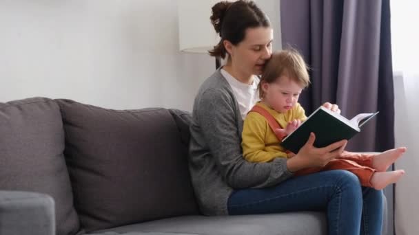 照看小孩的年轻妈妈抱着快乐可爱的小女孩抱着学习读书童话故事与家人团聚在家里享受家庭生活的乐趣坐在客厅里舒适的沙发上 — 图库视频影像