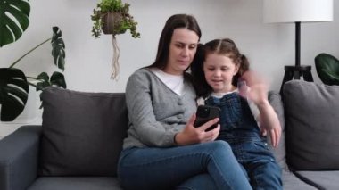 Mutlu genç anne ve küçük kızı akıllı telefondan video görüşmesi yapıyor akrabalarıyla konuşuyor, gülümseyen kız ve anne cep telefonlarına el sallıyor, evden sohbet ediyorlar. Çocuklar ve teknoloji konsepti