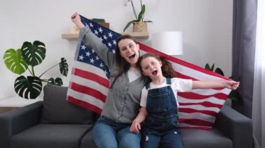 Neşeli genç bir anne ve gülümseyen küçük bir kız çocuğu evde rahat bir kanepede oturmuş Amerika 'nın büyük bayraklarıyla Bağımsızlık Günü' nü kutlarken kameraya bakıyor. Vatansever Amerikan bayramı konsepti