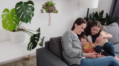 Gülümseyen genç anne ve iki tatlı kızın evdeki rahat koltukta oturup akıllı telefondan komik videolar izlemesi. Mutlu anne ve çocuklar hafta sonları cep telefonunda çizgi film seyrediyor.