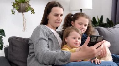 Neşeli anne ve iki küçük kız cep telefonuyla selfie çekiyorlar, video görüşmesinde konuşuyorlar, oturma odasında rahat bir kanepede dinleniyorlar, online medya izliyorlar, internetten alışveriş uygulaması kullanıyorlar.