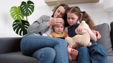 Sevgi dolu anne ve iki kızı akıllı telefondan komik videolar izleyip rahat bir oturma odasında rahat koltukta oturuyorlar. Gülümseyen anne ve çocuklar hafta sonları cep telefonunda çizgi film seyrediyorlar.