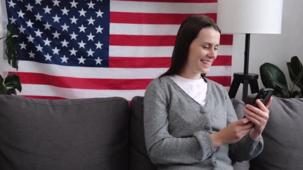 Sonriente Joven Americana Sentada Cómodo Sofá Casa Con Bandera Agitando — Vídeo de stock