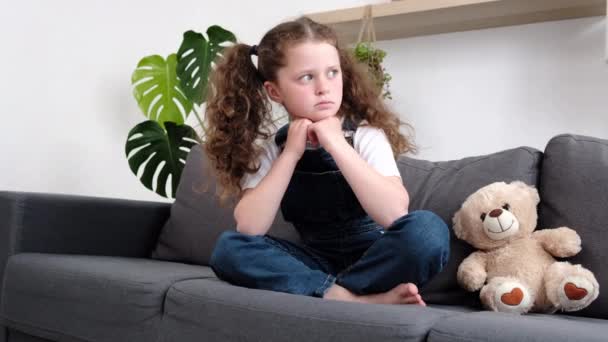 心烦意乱的 被欺负的小女孩的形象让人感觉被抛弃了 悲伤的 不幸的学童孤儿一个人坐在玩具玩具玩具旁的灰色沙发上 慈善收养的概念 — 图库视频影像