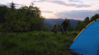 Aile aktivitesi macera konsepti. Sarı bereli genç bir baba ve gün batımında bulutlu gökyüzünde dağlarda yürüyen bir kız çocuğu. Babası ve şapkalı kızı yaz doğasının tadını çıkarıyorlar.