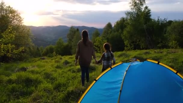 キャンプで夕方に夕日を見ている若い母親と小さな娘の子供のバックビュー 山の背景を歩いて小さな女の子の子供とお母さんの世話をする 休暇のコンセプトの家族活動の冒険 — ストック動画
