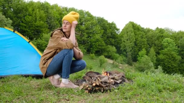 一个身披黄色大礼帽 坐在篝火边和高山背景上蓝色帐篷里的快乐而浪漫的年轻女子的画像 冷静的女孩喜欢大自然 周末出游 宁静和专注的概念 — 图库视频影像