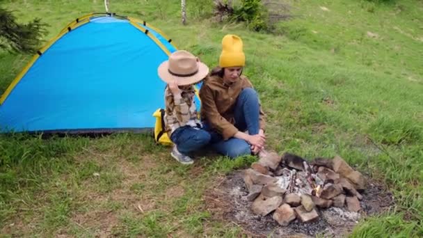 年轻的母亲和戴着帽子的可爱的小女儿喜欢坐在篝火和帐篷边露营 妈妈和小女孩一起过周末 度假家庭活动探险概念 — 图库视频影像
