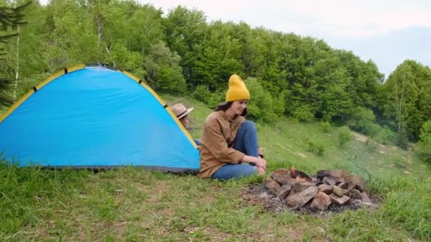 快乐的年轻妈妈 身穿黄色的针织便帽 可爱的小女儿 坐在靠近蓝色帐篷和篝火的山顶上 享受美丽的大自然 流浪和旅游概念 — 图库视频影像