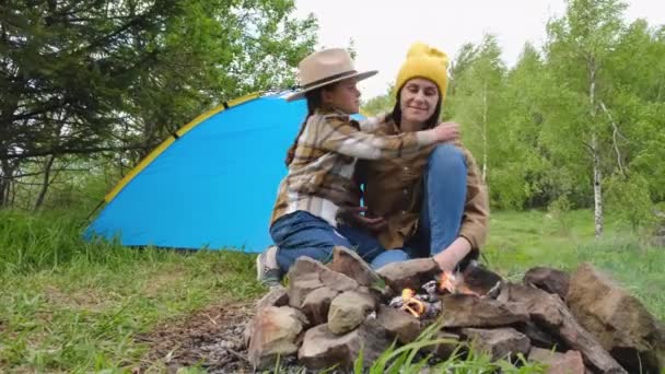 快乐的年轻妈妈和可爱的小女儿戴着帽子坐在篝火边的绿色森林里 笑得漂亮的小孩抱着妈妈 享受夏天的自然 和孩子一起在大自然中露营 — 图库视频影像