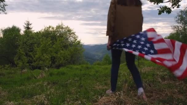 戴着礼帽的美丽年轻的高加索女人 独立而年轻 站在多云的天空和夏日的群山中 高高地高举美国国旗 体现了爱国心和自由 — 图库视频影像