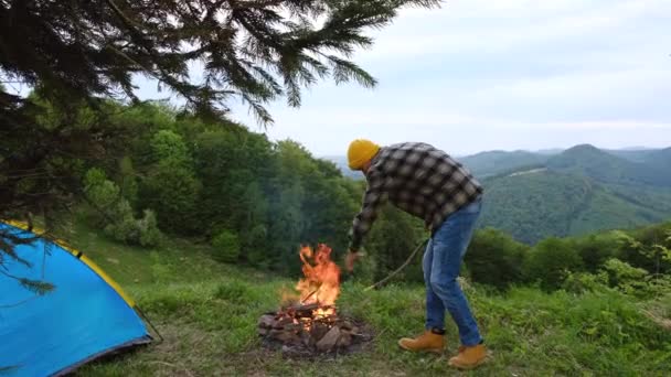 若いひげを生やした男は緑の森と美しい夏の山々を眺めながらテントの近くの丘の上に座って ヒップスター男は火を燃やす キャンプライフ 都市から脱出する 観光の概念 — ストック動画