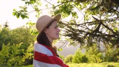 Amerikan bayrağına sarılmış bej şapkalı güzel beyaz kadın portresi. Günbatımında ya da gün doğumunda doğanın tadını çıkaran hippi tebessümlü kıza yakın çekim. 4 Temmuz, Bağımsızlık Günü konsepti