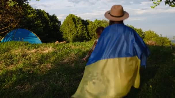 年轻的乌克兰母亲头戴礼帽 带着小女儿独立站在背景的夏山上 自豪地高举着乌克兰国旗 冒险和自由概念 — 图库视频影像