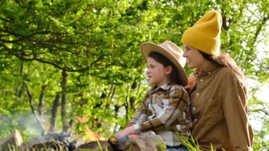 Arka plandaki yeşil ormanda şenlik ateşinin yanında oturan mutlu anne ve küçük kız. Gülümseyen annesi olan sevimli küçük çocuk yaz doğasının tadını çıkarıyor. Açık hava kampı yaşam tarzı, şehir dışı göçebeler.