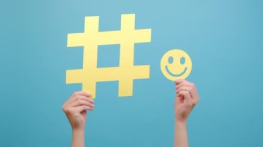 Büyük sarı hashtag işareti ve gülümseyen küçük bir yüz ikonu tutan kadınların ellerini kapatın. Stüdyonun mavi arkaplan duvarında reklam için fotokopi alanı olan izole edilmiş. Çevrimiçi ve web kavramı