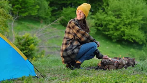 夕阳西下 戴着黄色帽子的快乐的年轻女性坐在篝火边 蓝色的帐篷在夏山的背景下 沉着微笑的女孩喜欢大自然 周末出游 宁静和专注的概念 — 图库视频影像
