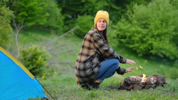 漂亮的年轻女子坐在篝火边 帐篷在背景森林里 穿着黄色贝尼的迷人的快乐女孩喜欢夏天的自然生活 周末出游 宁静和专注的概念 慢动作 — 图库视频影像