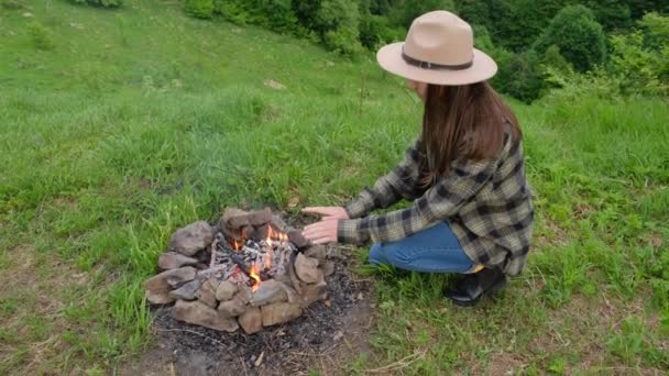 活跃的年轻女子头戴礼帽坐在篝火边 在背景夏季的森林里燃烧着 迷人的黑发女孩20多岁喜欢大自然 周末出游 宁静和专注的概念 — 图库视频影像