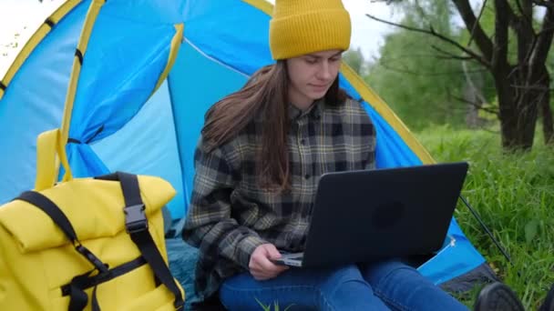 在户外的蓝色帐篷里 坐在笔记本电脑上工作的年轻女人被夏日的自然包围着 在假期里远程工作的女性自由职业者很可爱 现代千年工作方式概念 — 图库视频影像