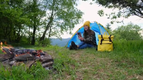 身穿针织黄帽和衬衫的快乐的年轻女子坐在帐篷里 用智能手机通过视频通话 并在户外用手拿着相机挥手 概念探险旅行博客Wanderlust — 图库视频影像