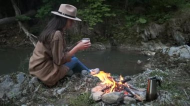 Şirin genç bayan gezgin, kamp ateşi ve dağ nehrinin yanında oturup çay ya da kahve içiyor. Kamp alanı atmosferi, doğada yaşam tarzı. Şehir yakınlarında bir gecelik kamp gezisi. Doğa manzaralı kamp alanı.