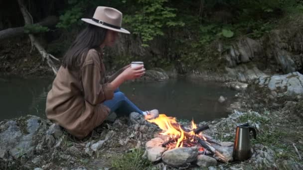 可爱的年轻女性旅行者坐在篝火边和山河边喝茶或喝咖啡 露营地的室外氛围 自然界的生活方式 城市附近的夜间露营旅行 自然风景营地 — 图库视频影像