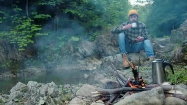 Örgü bereli genç adamın seçici odak noktası akşam vakti kamp ateşinin yanında sıcak kahve içmek taşın üzerinde oturmak. İnsanlar yaz aylarında şenlik ateşine bakar ve doğada çay içer. Macera ve seyahat konsepti