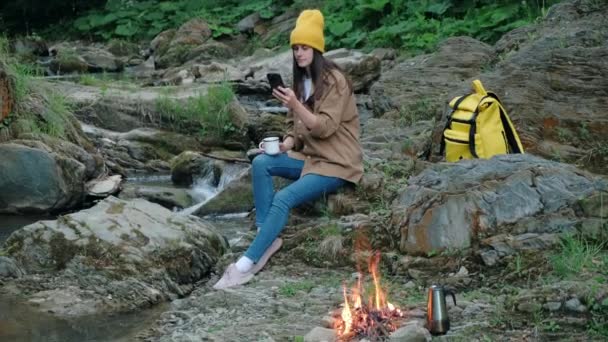 用智能手机徒步旅行的年轻女子 坐在山河边的石头上喝着热咖啡和篝火 美丽和鼓舞人心的营地位置 为室外生活的生活方式生动 旅行概念 — 图库视频影像