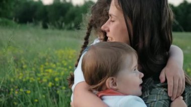 Aile mutluluğu, duygu konsepti. Mutlu gülümseyen genç anne, iki tatlı kızı kucaklıyor. Sakin bir yaz gününde yeşil çimlerde oturan çocuklar, sıcak anne hisleri, yaşam tarzı anı.
