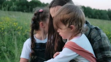 Gülümseyen genç anne ve iki tatlı kız kardeşin yan görüntüsü. Kucaklaşan, oynayan ve dışarıda eğlenen iki kız kardeş. Mutlu anne ve küçük güzel kız akşam vakti sakin doğanın tadını çıkarıyorlar.