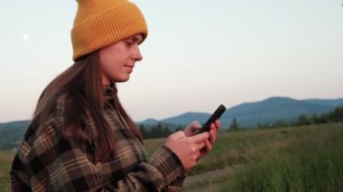 Sarı bereli şirin gülümseyen beyaz kadın, gün batımında dağ yürüyüşü sırasında internetten mesajlaşma ve internette gezinme içeriği. Seyahat kavramı