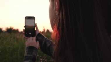 Selective focus of beautiful young woman 20s using smartphone, record video or make photos of amazing sunset and landscape scenery. Concept adventure travel blogger. Outdoors living lifestyle