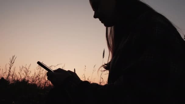 Силуэт Молодой Женщины Сидящей Траве Держащей Мобильный Телефон Время Заката — стоковое видео